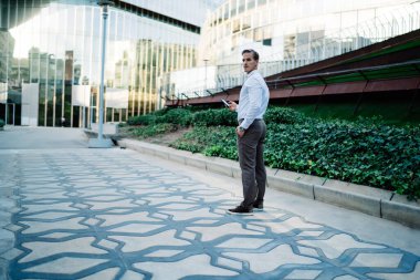 Modern mobil cihazlı akıllı girişimcinin tam boy portresi finans bölgesinde şehir merkezinde duruyor ve kameraya bakıyor, beyaz iş adamı şehir merkezinde poz veriyor.