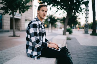 Şehir caddesinde dizüstü bilgisayarda yazı yazan Kafkas erkek blogcu, kameraya bakan dalgın adam boş zamanlarında sosyal ağlarda multimedya içeriğini paylaşıyor.