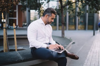 Olgun erkek tüccar online iş için dışarıda şehir ortamında oturuyor, orta yaşlı girişimci modern dizüstü bilgisayardaki web dosyalarını yüklemek için 4G kablosuz internet bağlantısı kullanıyor