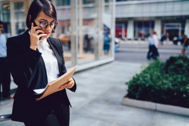 Resmi kıyafet ve gözlüklü ciddi bir iş kadını belgelerle uğraşırken ve New York 'ta sokakta dururken akıllı telefondan konuşuyor.
