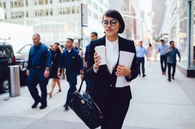Gözlüklü, düşünceli, genç bir ofis çalışanı. Resmi olarak elinde yemek fincanı ve belgelerle geride insanlarla birlikte kaldırımda yürüyor.