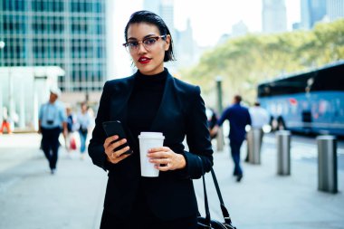 Başarılı, siyah saçlı, şık takım elbiseli, elinde kahve fincanı ve cep telefonu tutan şık gözlüklü bir kadın.