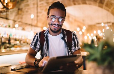 Gözlüklü, gülümseyen Afro-Amerikan hippi adam tablet üzerine kitap okuyor, kafenin içinde boş zaman geçiriyor, neşeli koyu tenli erkek 20 'li yaşlarda sosyal ağlarda bilgi aramaktan keyif alıyor.