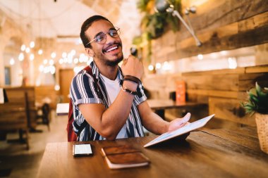 Neşeli etnik erkek serbest giyinmiş ve gözlüklü, akıllı telefon ve defterle kafede parlak ışıklarla otururken tablet kullanıyor.