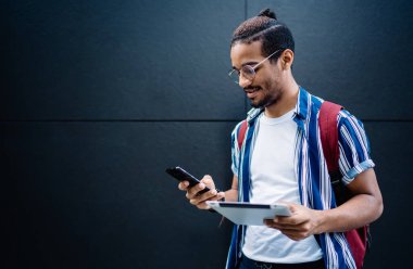 Koyu tenli hippi 4G bilgilerine bağlı dijital tablet tutarken akıllı telefondan numara çeviriyor. Genç Afro-Amerikan erkek, şehir duvarında duran mesajlar ve mesajlar gönderiyor.