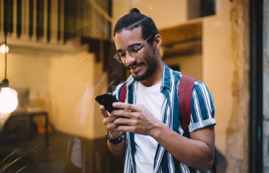 Günümüz modern binasının cam duvarında dikilirken sırt çantalı ve sırt çantalı, kendine güvenen siyah saçlı bir adam akıllı telefonuyla sörf yapıyor.