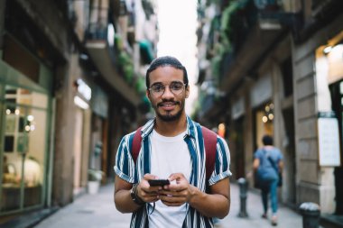 Neşeli, sakallı, sıradan kıyafetler ve gözlüklü bir etnik adam yaya caddesinde duruyor ve kameraya bakıyor.