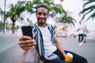 Mutlu, sakallı, sıradan giyinmiş, bardaklı, tek kullanımlık kahve bardağıyla bankta otururken selfie çeken etnik erkek.