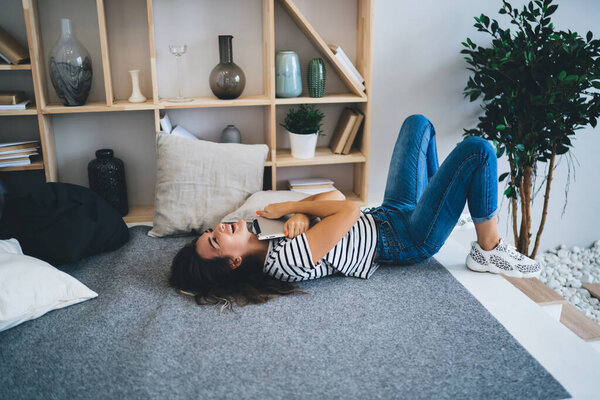 High angle of happy excited young brunette laughing and hugging tablet while lying on floor in modern apartment after getting good news