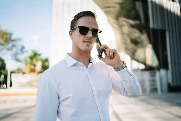 Serious male employee in formal shirt and sunglasses communicating on phone while standing on street near modern building on sunny day