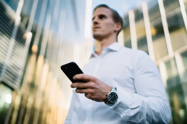 Below view on blurred Caucasian businessman with wearable swiss watch on hand holding modern smartphone device and thinking in financial district, male entrepreneur using mobile gadget in downtown