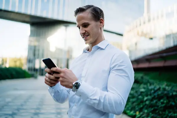 Successful employee enjoying cellular chatting standing at metropolitan urbanity in downtown and using 4g wireless connection, happy Caucasian businessman installing banking application and smiling
