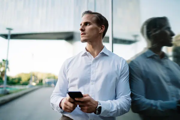 Contemplative male in white shirt standing near urban building thinking about financial news received on smartphone device, millennial businessman using cellular technology in metropolitan downtown