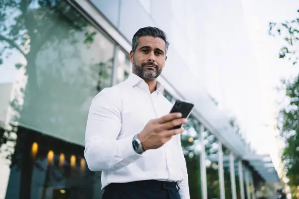 Formally dressed businessman with mobile phone standing at urban setting of financial district and looking at camera while using phone tech, half length of mature Caucasian employer with smartphone