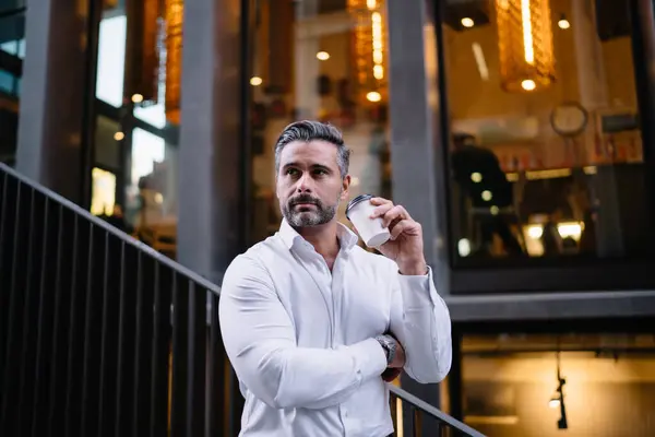 Pensive Caucasian businessman in white shirt holding caffeine beverage in disposable takeaway cup standing at urban setting and thinking in downtown, middle aged male employer with coffee to go