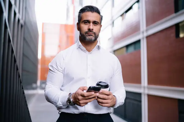 Half length portrait of confident proud CEO looking at camera during coffee break in city, Caucasian middle aged corporate director with cellular device and takeaway cup posing at urban setting