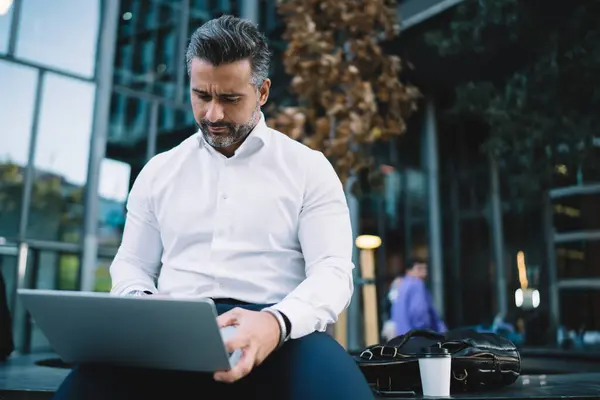 Skilled businessman in white shirt puzzled on received laptop information during remote work in financial district, Caucasian male 40s making online booking and banking via netbook technology