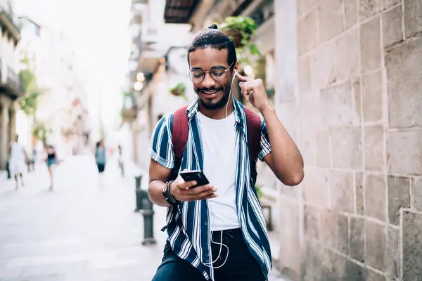Positive dark skinned hipster guy meloman in earphones checking playlist on mobile phone walking on street, smiling african american tourist enjoying online radio via 4G internet connection in roaming