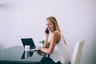 Fotoğraf makinesine gülümseyen, telefon eden, netbook 'la parlak masasında oturan, beyaz duvarda beyaz ofis duvarına karşı sıcak bir bardak içki içen neşeli sarışın kadının yan görüntüsü.