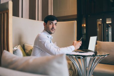 Lobide dizüstü bilgisayarla oturup kameraya bakarken interneti cep telefonuyla gezen kararlı etnik erkek girişimci.