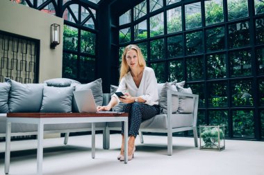 Resmi kıyafetler giymiş, netbook kullanan ve kafede oturup kameraya bakarken akıllı telefondan sörf yapan kadın bir patron.