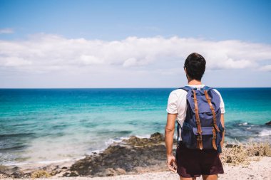 Sırt çantalı genç erkek turistin kıyı şeridinde durup deniz manzarasını izlerken dinlenen ve sakin denizin tadını çıkaran görüntüsü.