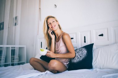 Mutlu genç kız sabah kahve içerken, rahat bir dairede, çay fincanı taşıyan neşeli beyaz kadın cep telefonu yapmak için 4G kablosuz telefon kullanıyor.