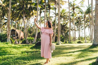 Tam boy, pozitif, genç bir kadın arkada şık pembe Maxi elbiseler giyiyor çimenli çayırlarda duruyor ve tropikal tatil beldesinde cep telefonundan selfie çekiyor.