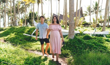 Tam vücut iyimser genç hamile kadın şık pembe elbiseler içinde mutlu kocayla el ele tutuşurken egzotik kır bahçesinde ahşap kulübelere karşı birlikte yürüyorlar.