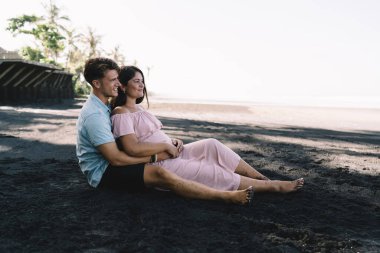 Çok ırklı, çıplak ayaklı, pozitif bir çiftin kumsalda, küçük bir yerleşim yerinin yanında oturması ve yaz günü denizin tadını çıkarması.