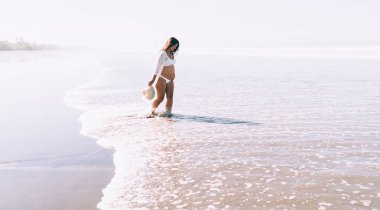Bikinili ve plajlı hamile bir kadın, kumlu sahilde gün ışığında dalgalı deniz suyunda yürürken şapka takıyor.