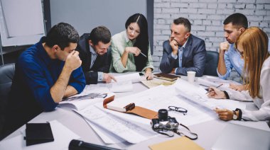 Group of male and female colleagues collaborating on blueprint and design project discussing strategy indoors, experienced architects experts talking about marketing plan and paper sketches