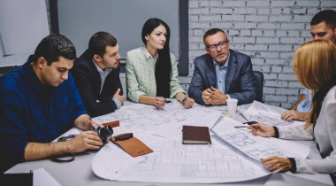 Group of male and female colleagues collaborating on blueprint and design project discussing strategy indoors, experienced architects experts talking about marketing plan and paper sketches