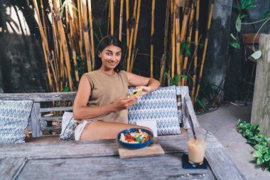 Sıradan giyinen, gülümseyen ve kameraya bakan pozitif etnik kadın elinde akıllı telefonuyla tropikal kafe bankında dinleniyor.