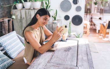Ahşap masadaki rahat kafeteryada taze meyve suyu bardağının yanında oturmuş fotoğraf çeken içerikli etnik kadın manzarası