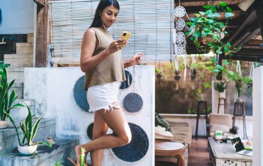 Bacağı kalkmış, verandada saksı bitkileriyle duran ve kulaklıkla müzik dinleyen mutlu bir etnik kadın.