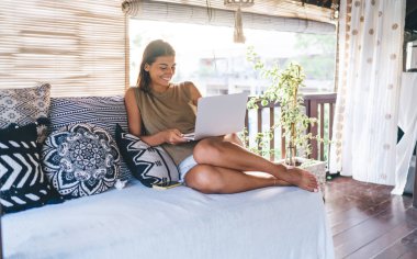 Bali tatil köyünde dizüstü bilgisayar kullanırken ferah ışık odasında rahat koltukta tek başına oturan çıplak ayaklı, etnik bir kadın.