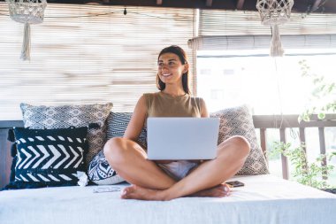 Keyifli, koyu renk saçlı, yaz kıyafetleri içinde, Lotus pozunda dizüstü bilgisayarla yastıklı koltukta oturan genç bir kadın.