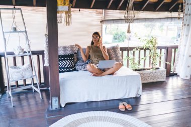 Gözlüklü genç bayan rahat koltukta dizüstü bilgisayarıyla oturuyor ve güneşli bir günde cep telefonuyla konuşurken el kol hareketi yapıyor.