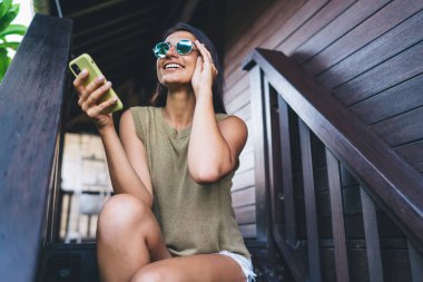 Neşeli, etnik bir kadının güneş gözlüğünü ayarlaması ve tropik tatil köyünde cep telefonuyla sörf yaparken terasta ahşap merdivenlerde oturması.