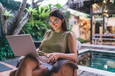 Haftasonu dinlenirken dizüstü bilgisayarı karıştıran pozitif etnik bir kadın ve avluda bacak bacak bacak üstüne atmış rahat bir koltukta oturuyor.