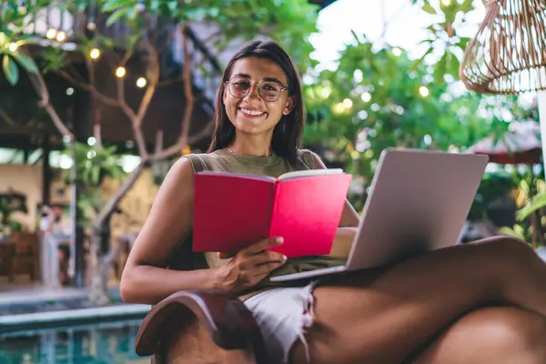 夏休み中にプールサイドに座ってノートパソコンやノートブックを使った宿題をしながら 陽気な若いエスニック女性学生が笑顔で ストック画像