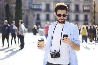 Sıradan giyinmiş, güneş gözlüklü, sokakta fotoğraf makinesi olan ve akıllı telefondan kahve ile mesajlaşan kendine güvenen sakallı bir adam.