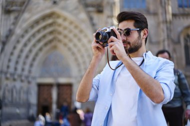 Sıradan kıyafetleri ve güneş gözlükleriyle caddede dikilip eski binanın fotoğrafını çeken konsantre sakallı bir gezgin.