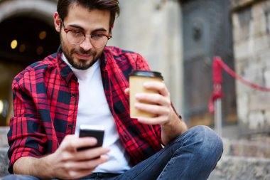 Sıradan giyinmiş, gözlüklü, sakallı bir adam cep telefonuna mesaj yazıyor ve dışarıda dinlenirken kahve içmenin tadını çıkarıyor.