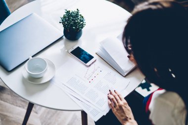 Akıllı kadın, grafik çizelgeleriyle masa başında dizüstü bilgisayar ve cep telefonuyla kağıt analizi yapıyor. Uzaktan çalışan profesyonel bir kadın, bilgileri kontrol ediyor ve bilgi okuyor.