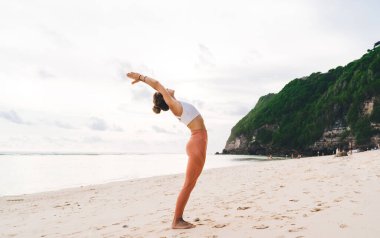 Bereketli yeşilliklerle kaplı kayalık kayalıkların yakınındaki kumlu sahile uzanmak için Urdhva Hastasana yapan esnek kadının yan görüntüsü