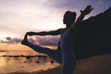 Gün batımında deniz kenarında yoga yapan genç ve formda bir kızın siluet görüntüsü. Esnek kadın vücut kaslarını germek için asana yapıyor. Doğada uyum ve farkındalık hissediyor.