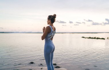Spor kıyafetleri içinde tanınmayan bir kadının deniz kıyısında yoga yaparken avuçlarını göğsüne yaklaştırıp akşam vakti deniz suyuna karşı meditasyon yaparken görüntüsü.