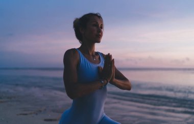 Spor elbiseli, genç, barışçıl etnik bayan sporcu gün batımı egzersizi sırasında okyanus kıyısında dua eden ellerle yoga yapıyor ve eğitim veriyor.